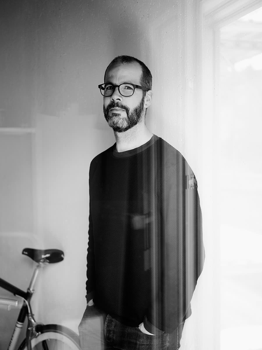 Schwarzweissfoto eines Mannes mittleren Alters mit Bart, kurzem Haar, schwarzem Sweater und gut gebrauchten Jeans, angelehnt an die weisse Wand seines Ateliers. Im Hintergrund ein extrem schönes Fahrrad – nicht drapiert im Fall.
